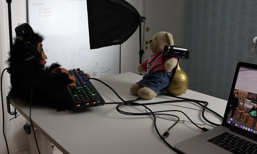 A chimpanzee types and wears headphones, while a teddy bear records him with a camcorder.